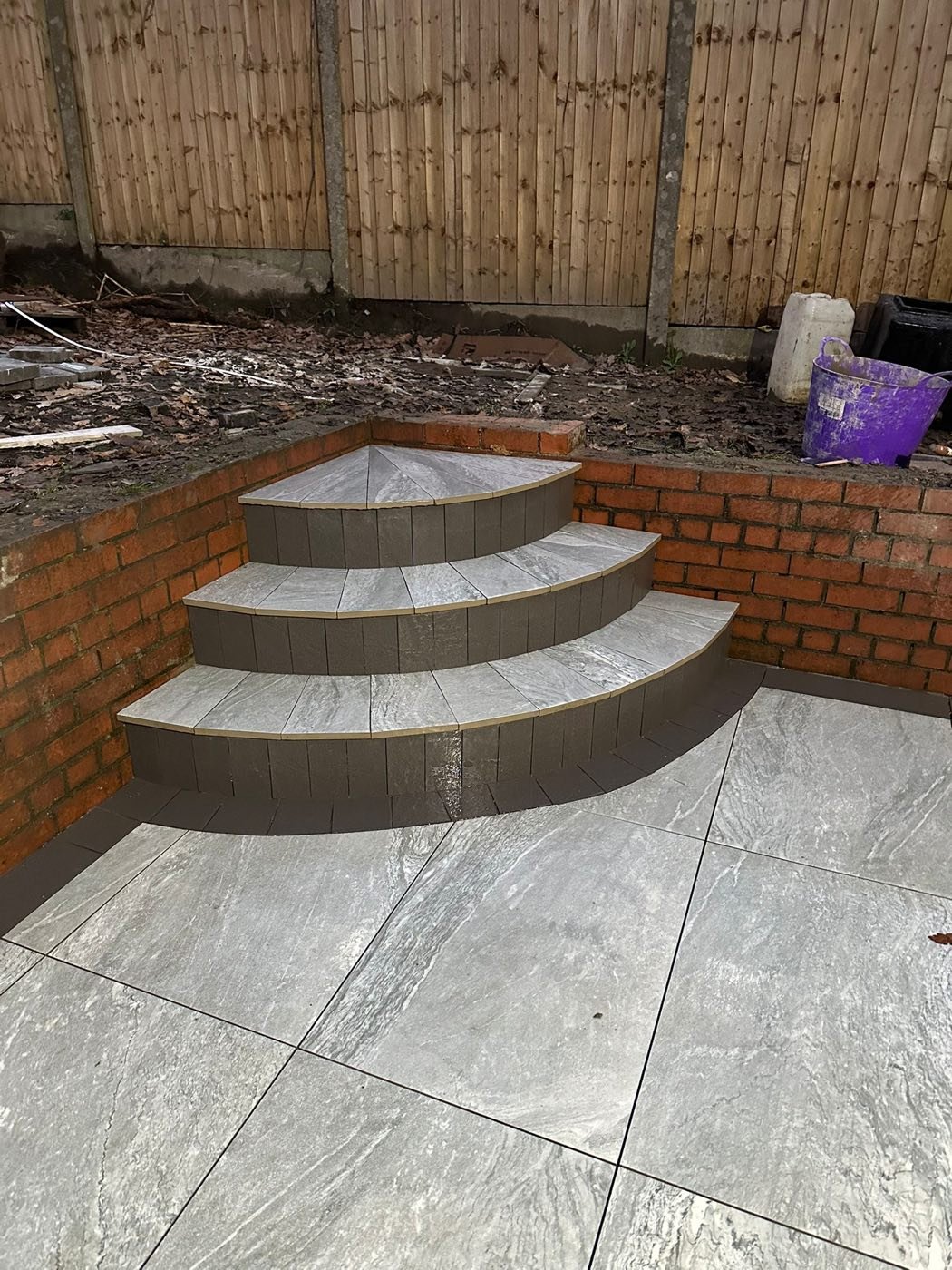 Curved three-tier grey porcelain corner steps with brick risers by North West Landscapes leading to a matching porcelain patio