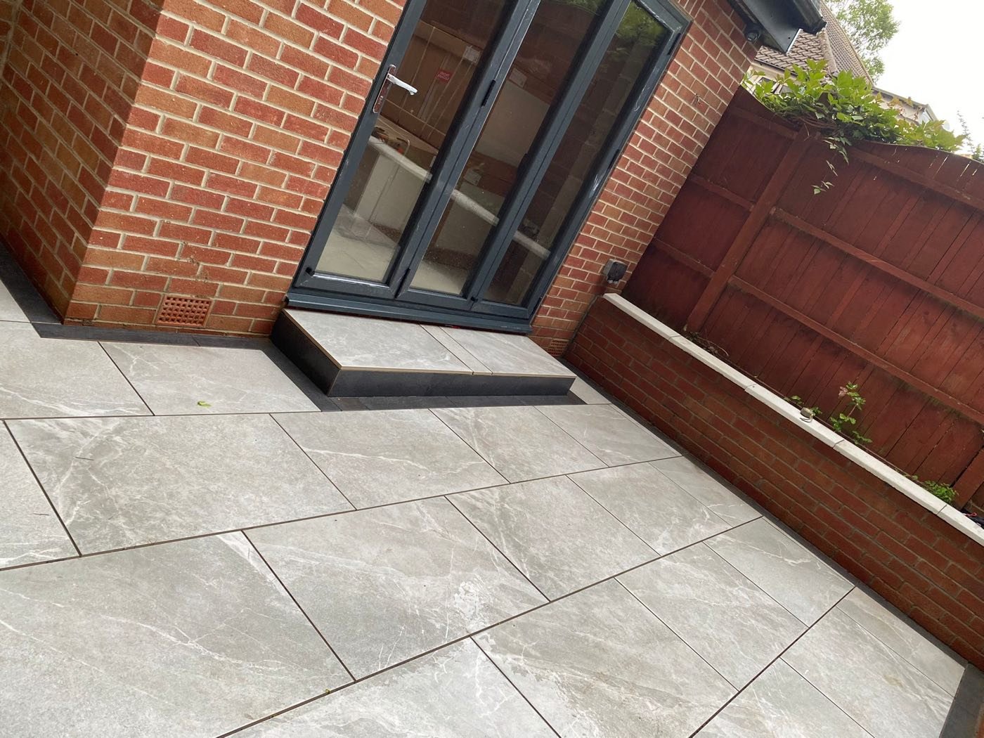 Grey marble-effect porcelain patio with charcoal step and anthracite doors in a back garden by North West Landscapes