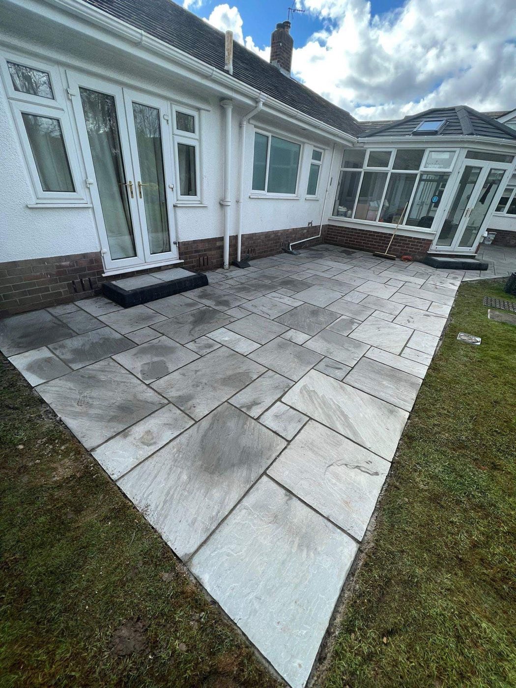 Large grey Indian sandstone patio by North West Landscapes at a rear bungalow garden with conservatory under blue sky