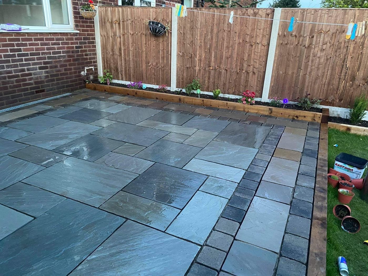 Grey Indian sandstone patio with charcoal block border and timber sleeper raised beds by North West Landscapes