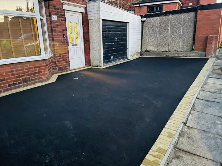 Finished black tarmac driveway with a buff block-paving border by North West Landscapes to a front of a red-brick semi