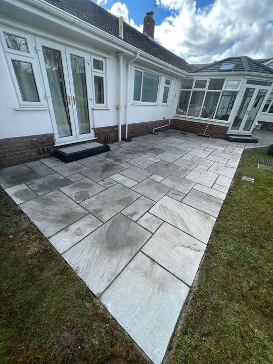 Large grey Indian sandstone patio by North West Landscapes at a rear bungalow garden with conservatory under blue sky