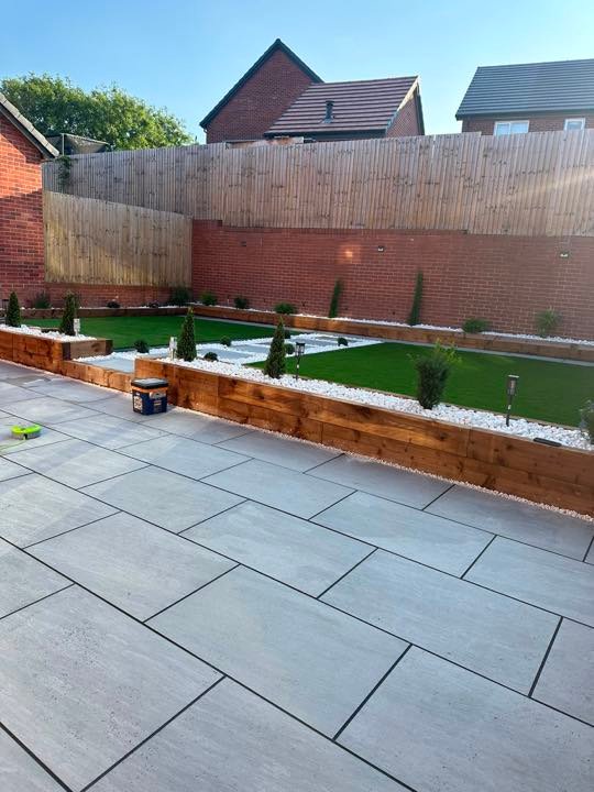 Large-format porcelain paving beside sleeper raised beds and artificial grass by North West Landscapes